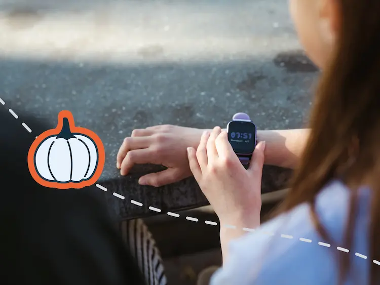 Kind schaut auf eine lilafarbene GPS-Kinderuhr am Handgelenk, daneben eine Halloween-Kürbisillustration.