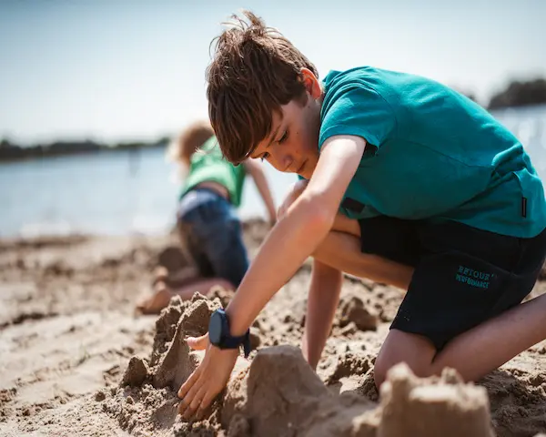 Kind mit GPS-Kinderuhr spielt am See und baut eine Sandburg.